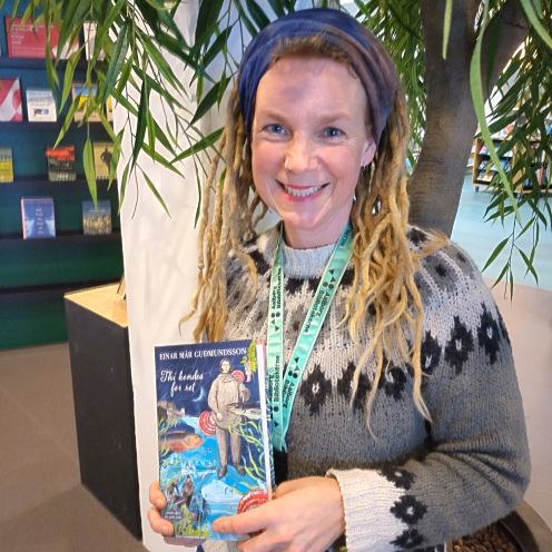 Karina står i afdelingen for skønlitteratur for voksne på Hovedbiblioteket. Hun holder "Thi kendes for ret" i hænderne og smiler til kameraet.