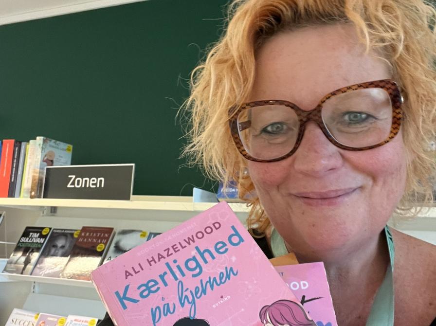 Maria står med bogen 'Kærlighed på hjernen' foran en zonereol i Hals.