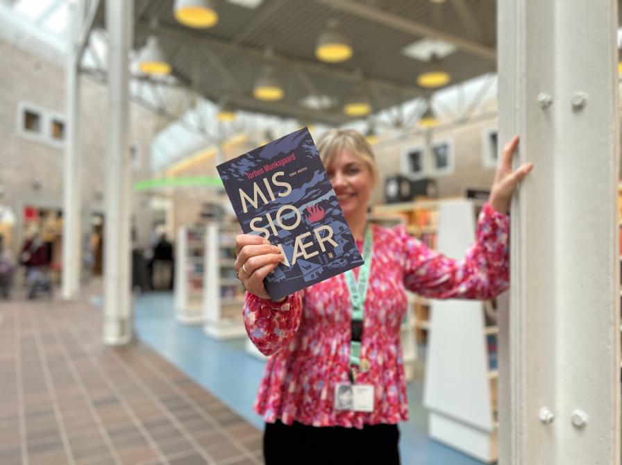 Marianne står på Hovedbiblioteket. Hun holder bogen "Missionær" frem for sig. Der er fokus på bogens forside, mens både Marianne og baggrunden, der viser biblioteksrummet, er let sløret. 