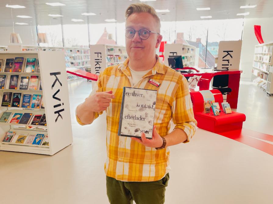 Martin står på Hjørring Bibliotek. Han holder den omtalte bog i denne artikel i den ene hånd og peger på den med den anden. Bag Martin ses biblioteksrummet med fyldte bogreoler og udstillinger.