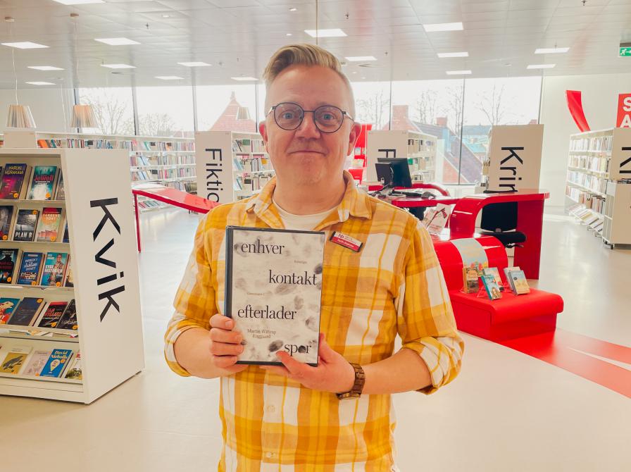 Martin står på Hjørring Bibliotek. Han holder den omtalte bog i denne artikel mellem hænderne. Bag Martin ses biblioteksrummet med fyldte bogreoler og udstillinger.